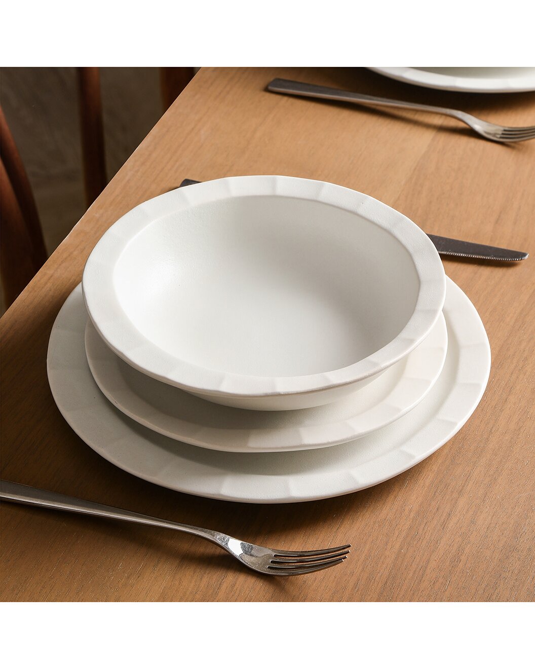 A place setting on a wooden table with a white bowl stacked on two white plates, accompanied by a fork and knife beside them.