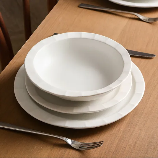 A place setting on a wooden table with a white bowl stacked on two white plates, accompanied by a fork and knife beside them.