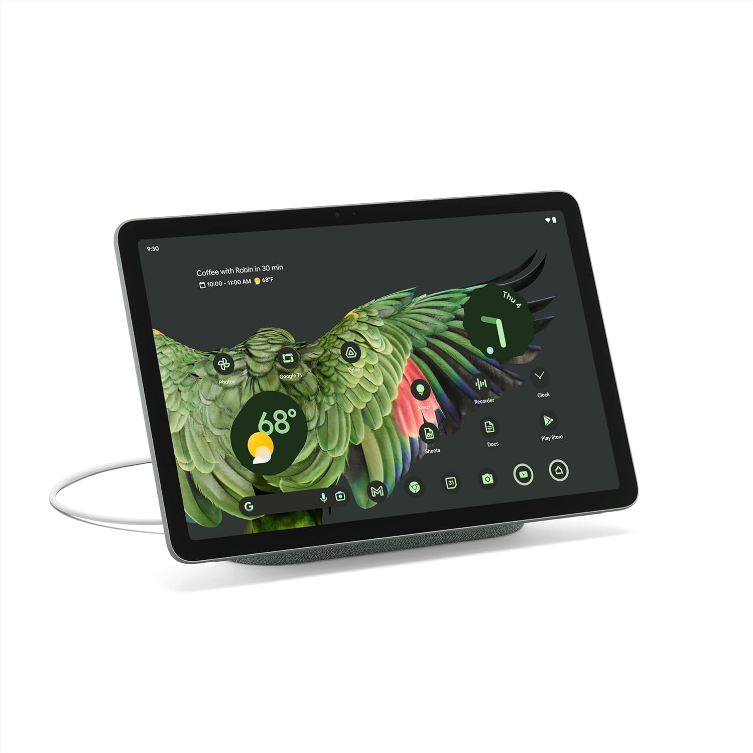 A Google Pixel Tablet mounted on a Speaker Dock, displaying a home screen with a green parrot-themed background and circular icons. The device is connected to a charger, which can also enhance audio output.