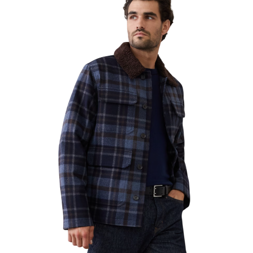 A man is wearing a blue and black plaid jacket with a brown collar, paired with a navy shirt and dark jeans.
