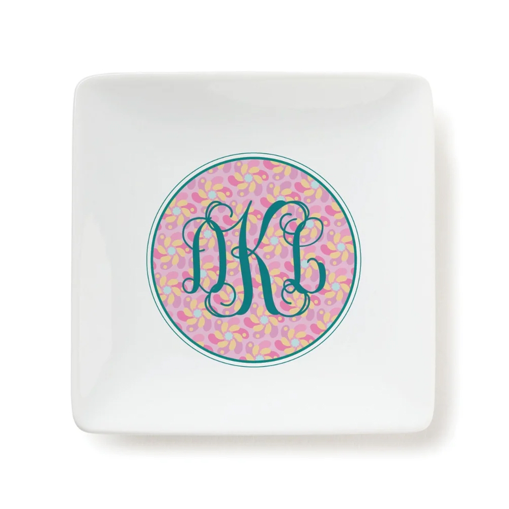 A square white trinket tray features a decorative circle with a pink and yellow abstract pattern and elegant teal monogram \“DKC\“ letters in the center.