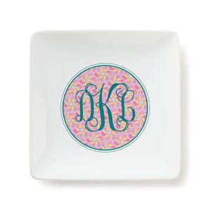 A square white trinket tray features a decorative circle with a pink and yellow abstract pattern and elegant teal monogram \“DKC\“ letters in the center.