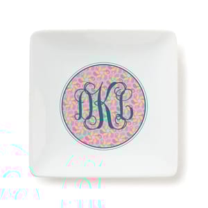 A square white trinket tray features a decorative circle with a pink and yellow abstract pattern and elegant teal monogram \“DKC\“ letters in the center.
