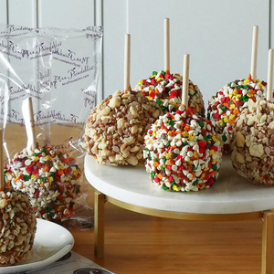 Caramel apples coated with chopped nuts and colorful sprinkle toppings are displayed on a white platter, with some individually wrapped in clear packaging labeled \“Mrs. Prindables.\“