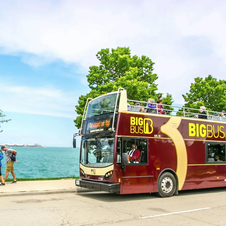 Double-decker sightseeing bus labeled \“Big Bus Chicago\“ drives alongside a body of water, with tourists sitting on the open top level and trees in the background.