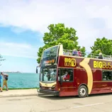 Double-decker sightseeing bus labeled \“Big Bus Chicago\“ drives alongside a body of water, with tourists sitting on the open top level and trees in the background.