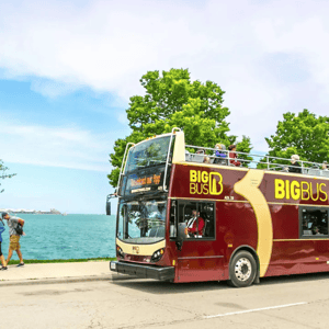 Double-decker sightseeing bus labeled \“Big Bus Chicago\“ drives alongside a body of water, with tourists sitting on the open top level and trees in the background.