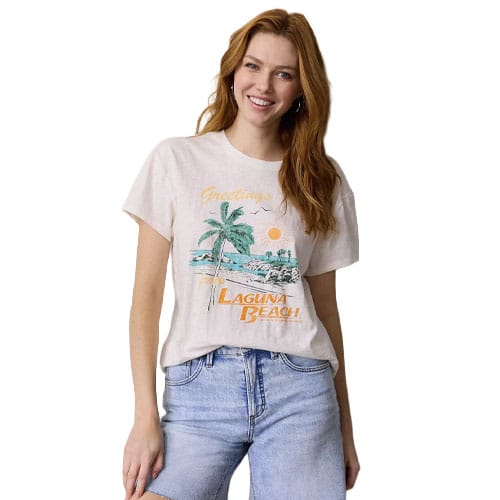 A woman is wearing a light-colored T-shirt with a \“Greetings from Laguna Beach\“ graphic featuring a palm tree and sunset, paired with light blue jeans.