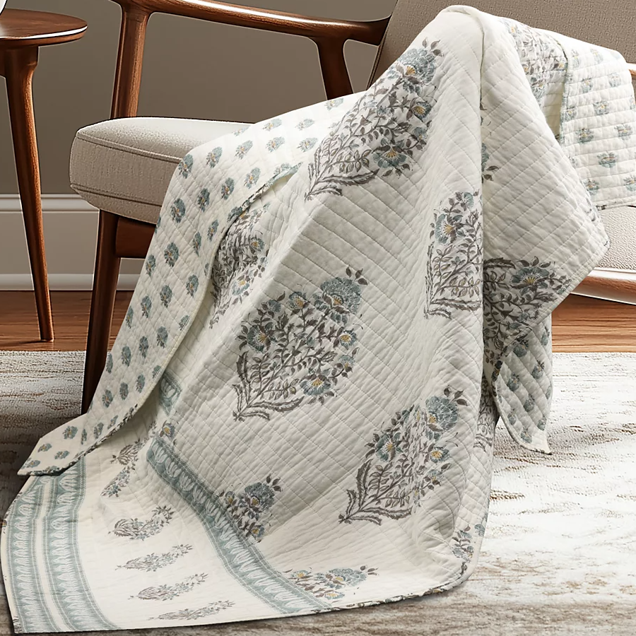 A quilt with a floral pattern in muted colors drapes over a beige upholstered chair, set on a light-colored rug in a room with wooden flooring.
