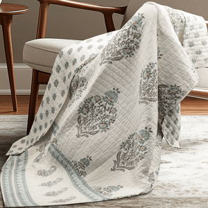 A quilt with a floral pattern in muted colors drapes over a beige upholstered chair, set on a light-colored rug in a room with wooden flooring.
