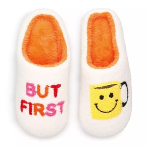 White slippers with fluffy orange interiors feature the words \“BUT FIRST\“ on one slipper and a smiling yellow coffee cup graphic on the other.