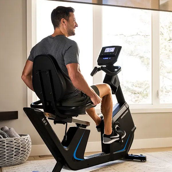 A man is exercising on a recumbent bike indoors, positioned by a large window, with a basket containing blankets nearby.