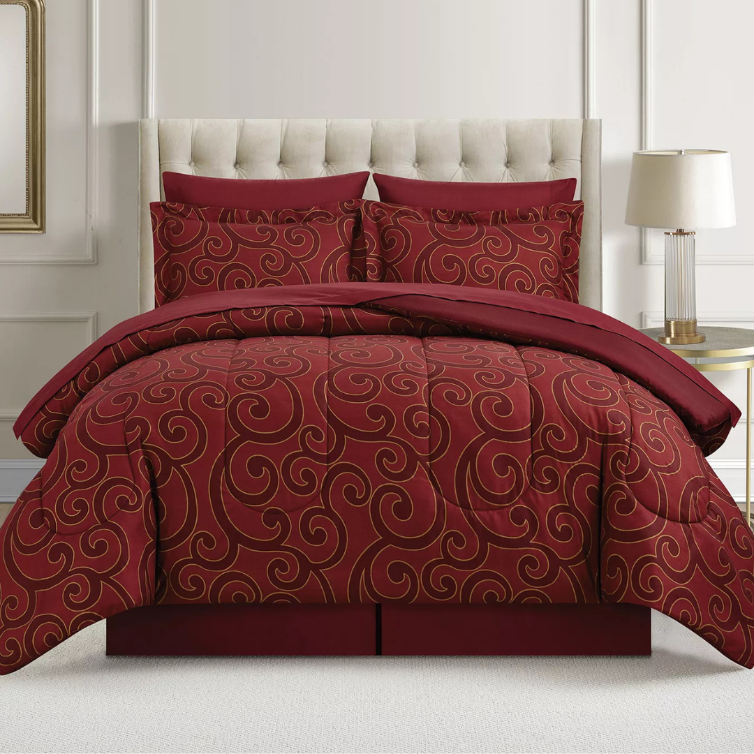 A red comforter set with swirling gold patterns is displayed on a bed with a tufted headboard. Two matching pillows are included, and a lamp on a side table is visible beside the bed.