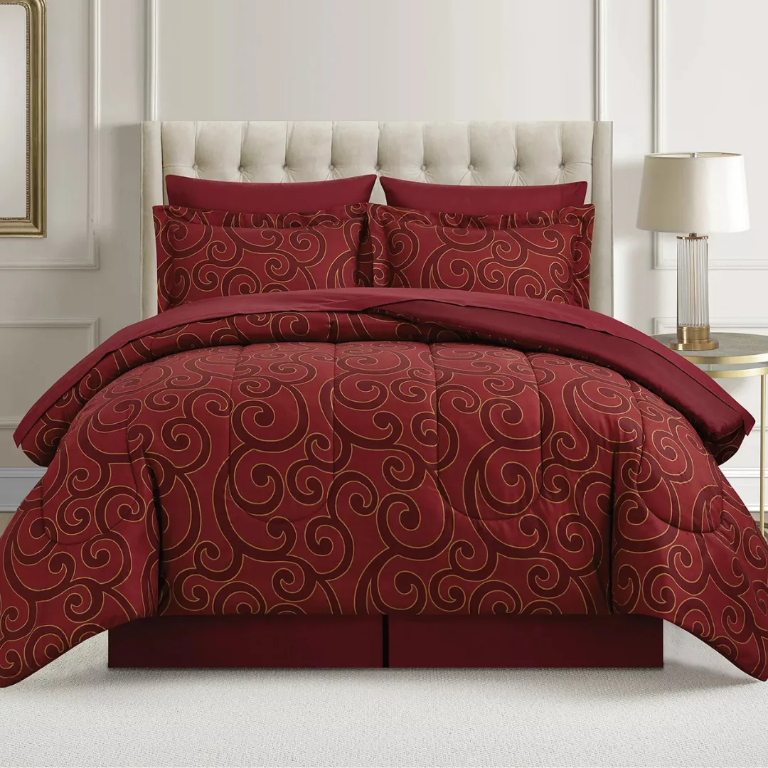 A red comforter set with swirling gold patterns is displayed on a bed with a tufted headboard. Two matching pillows are included, and a lamp on a side table is visible beside the bed.