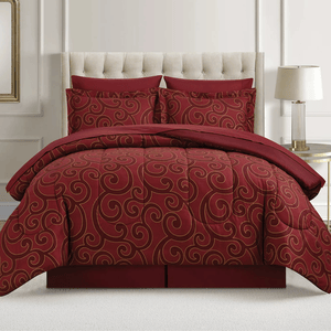A red comforter set with swirling gold patterns is displayed on a bed with a tufted headboard. Two matching pillows are included, and a lamp on a side table is visible beside the bed.
