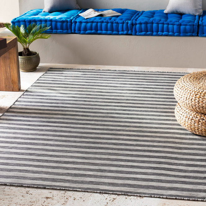 A striped rug in shades of gray and white is placed on a floor. It measures 8' x 10' and is suitable for both indoor and outdoor use. Nearby are textured poufs, a wooden table, and a cushioned bench.
