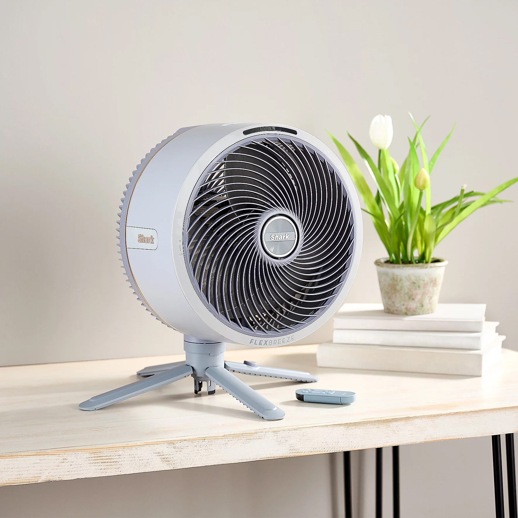A desk fan labeled \“Shark Flexbreeze\“ with a remote control sits on a wooden table next to a potted plant and stack of books.