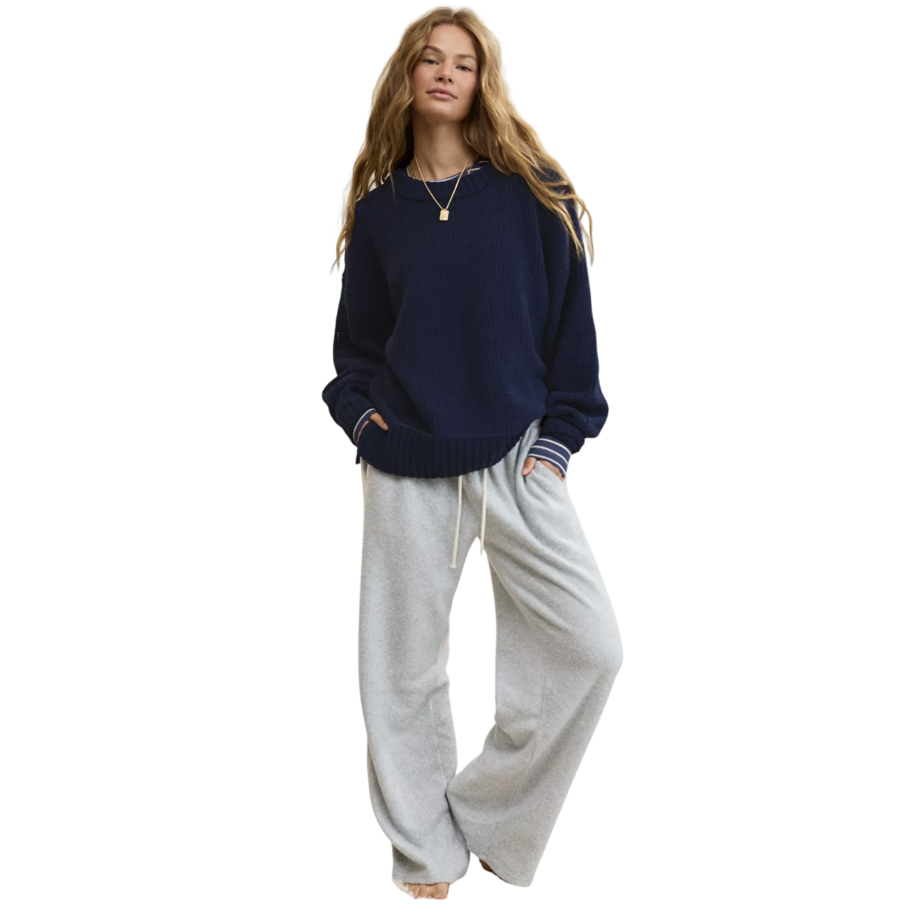 A person wearing a navy sweater, gray drawstring sweatpants, and layered necklaces stands on a cream carpeted floor.