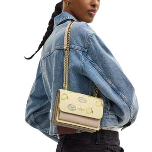 A person wears a denim jacket and carries a small yellow handbag with a chain strap, featuring a graphic design of sliced fruits.