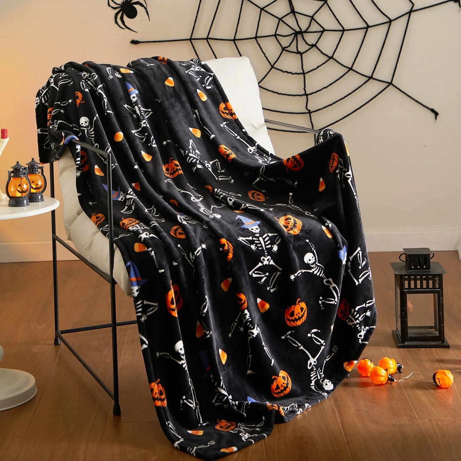 A Halloween-themed blanket with pumpkin, ghost, and skeleton prints draped over a chair, with a spider web decoration in the background.