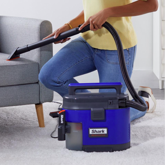 A person is using a Shark vacuum cleaner with a handheld nozzle to clean a couch. The vacuum is compact and has a purple and black design.