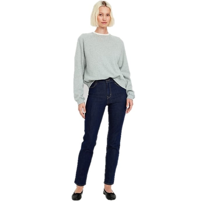 A woman is wearing dark blue, high-waisted straight-leg jeans that cut off at the ankle, paired with a light grey sweater and black flats.