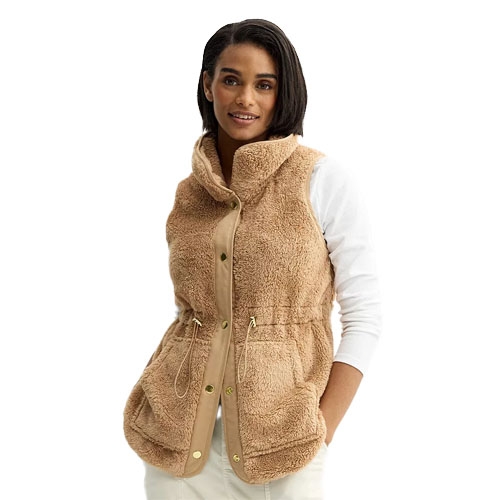 A woman is wearing a tan sherpa vest with a high collar and snap-button front, featuring a drawstring waist and large front pockets, paired with a white long-sleeve shirt.