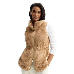 A woman is wearing a tan sherpa vest with a high collar and snap-button front, featuring a drawstring waist and large front pockets, paired with a white long-sleeve shirt.