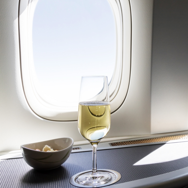A glass of white wine and a bowl of mixed nuts are on a table by an airplane window, illuminated by natural light.