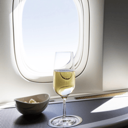 A glass of white wine and a bowl of mixed nuts are on a table by an airplane window, illuminated by natural light.