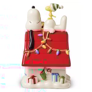 Lenox Snoopy & Woodstock Candy Jar features Snoopy lounging atop his red doghouse, adorned with colorful Christmas lights, and Woodstock wearing a green and white hat. The base is decorated with wrapped gifts, a candy cane, and a green stocking.