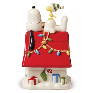Lenox Snoopy & Woodstock Candy Jar features Snoopy lounging atop his red doghouse, adorned with colorful Christmas lights, and Woodstock wearing a green and white hat. The base is decorated with wrapped gifts, a candy cane, and a green stocking.