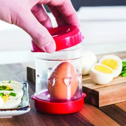 A hand is placing a lid on an egg peeler, which contains an unpeeled hard-boiled egg. Sliced eggs and chives are in the background on a cutting board.