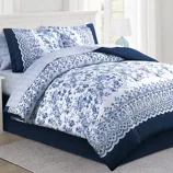 Blue and white floral-patterned bedding set with scalloped edges, including a comforter and matching pillow shams, on a bed with a white headboard.