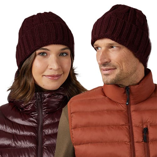 A burgundy cable knit beanie is shown, featuring a ribbed design for a snug fit, worn by a man and woman dressed in quilted jackets.