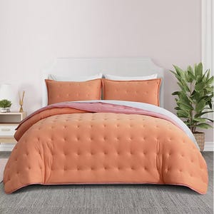 A peach-colored comforter set with matching pillow shams on a bed, accompanied by a plant and nightstand with decor items in a softly colored room.