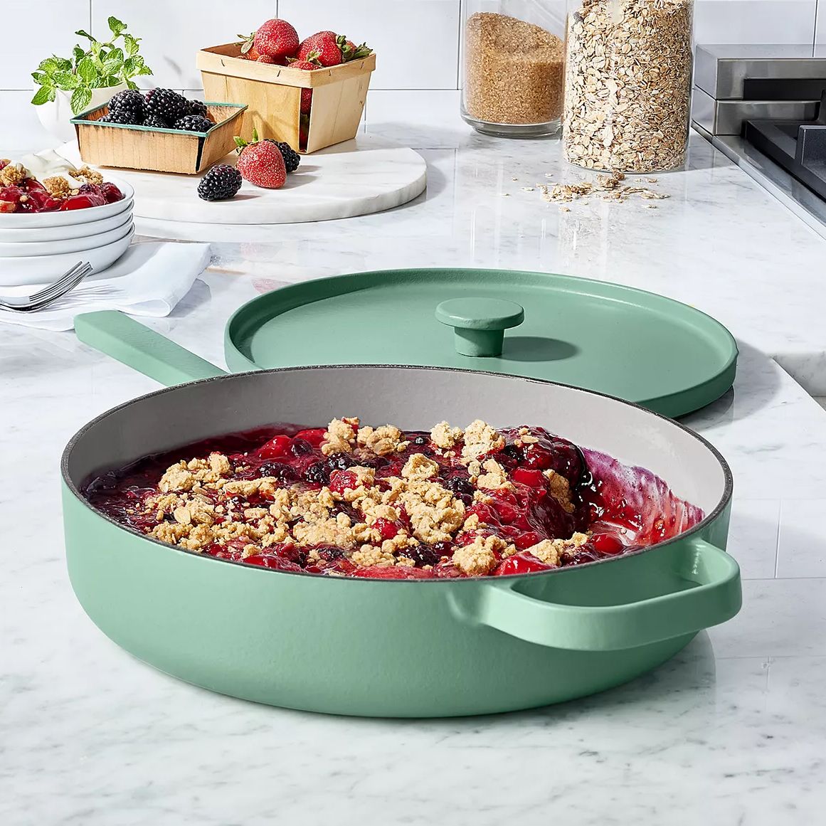 A green pan with a lid contains a berry crumble dish. Surrounding it are fresh berries, mint, plates with forks, brown sugar, and oats on a marble countertop.