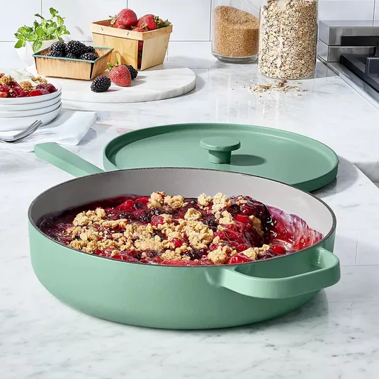 A green pan with a lid contains a berry crumble dish. Surrounding it are fresh berries, mint, plates with forks, brown sugar, and oats on a marble countertop.