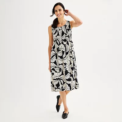 A woman is wearing a sleeveless, knee-length swing dress with a smocked bodice, featuring a black and white leaf print, paired with black flats.