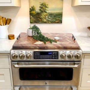 Wooden stove top cover with handles, featuring a herringbone pattern, placed on a stove. A small plant and ceramic dish are on top.