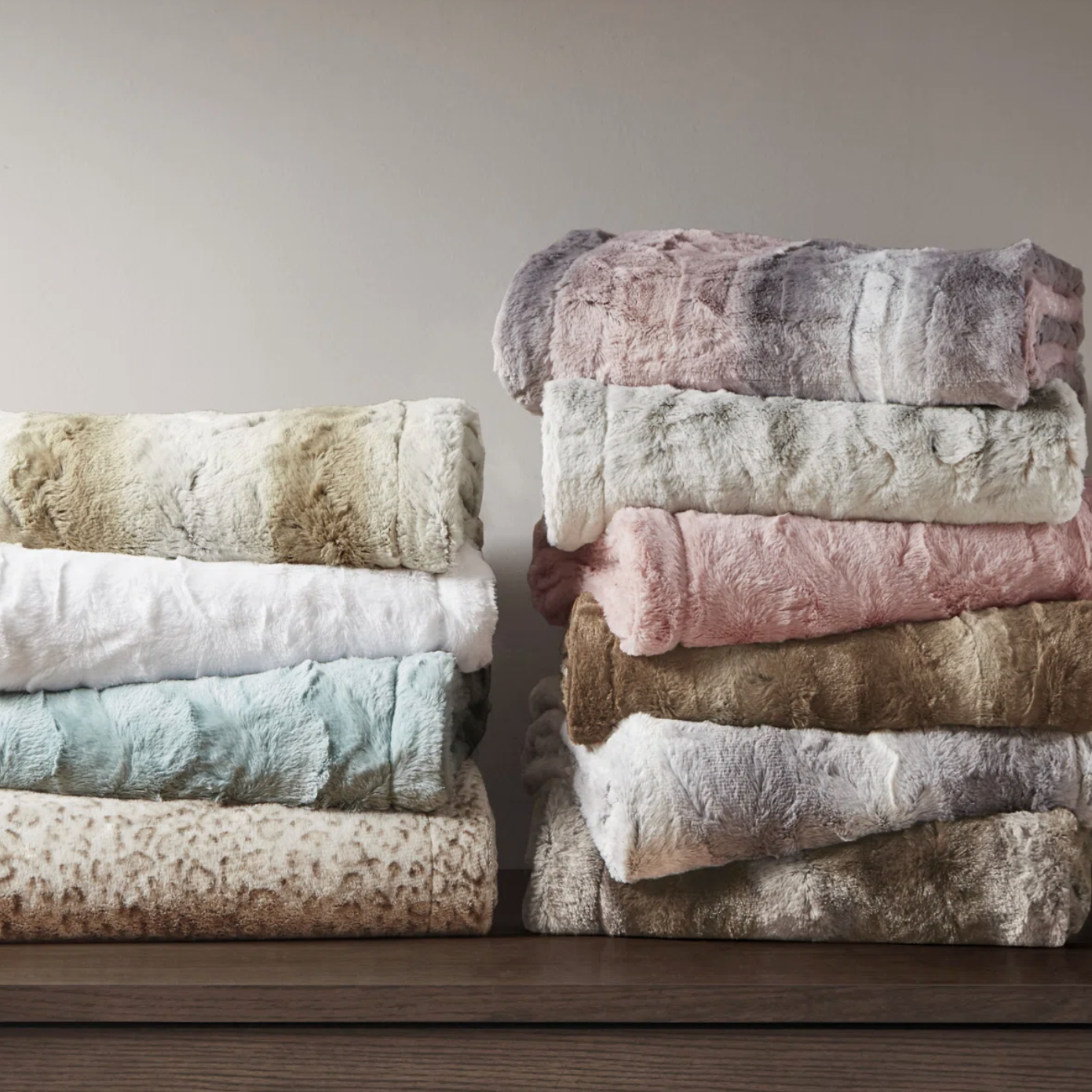 Two stacks of plush, faux fur throws in various colors, including white, pink, brown, and light blue, are neatly placed on a wooden surface.