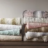Two stacks of plush, faux fur throws in various colors, including white, pink, brown, and light blue, are neatly placed on a wooden surface.