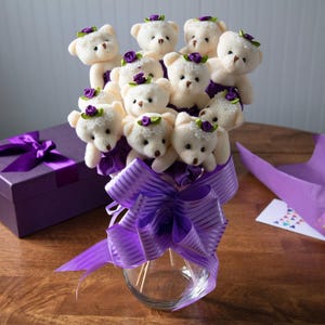 A bouquet of 12 small, plush teddy bears adorned with purple roses and beads, arranged in a clear vase with a large purple striped bow. Nearby is a purple gift box with a matching ribbon.