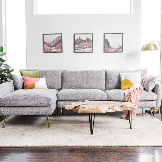 A gray sectional sofa with colorful pillows and a pink throw, accompanied by a natural wood coffee table with metal legs, is set on a textured rug. Three landscape paintings hang on the wall behind the sofa, and a gold floor lamp stands to the side.