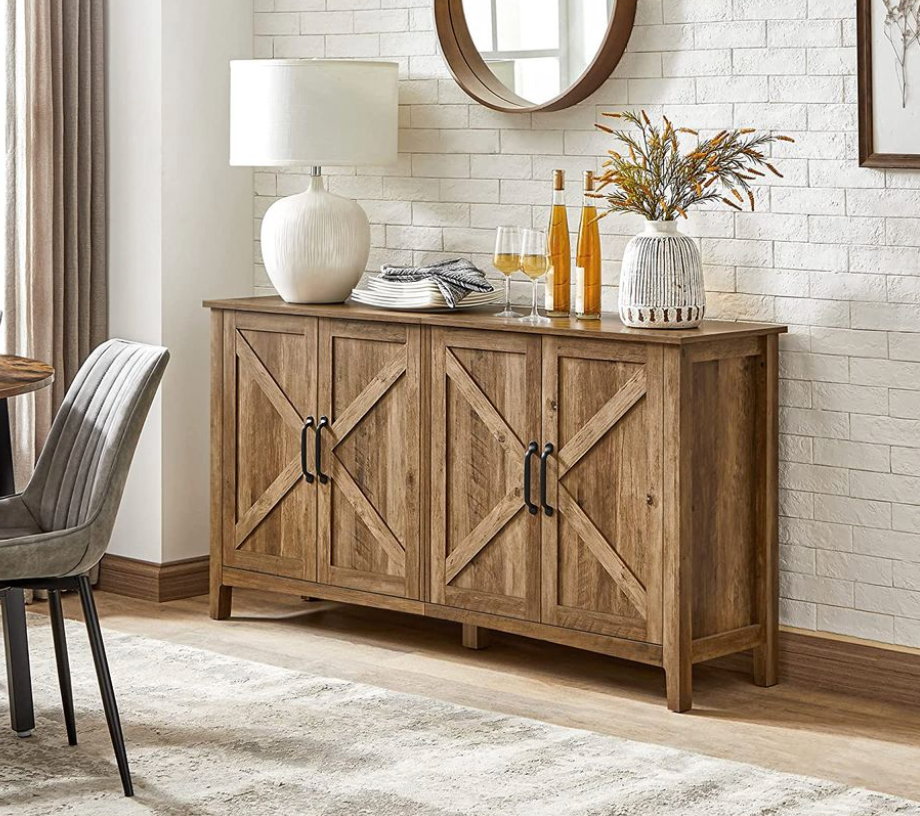 A rustic-style buffet cabinet with a wood finish and cross-patterned doors, featuring four handles. It is adorned with decorative items like a table lamp, plates, glasses, a vase, and bottles, set against a white brick wall.