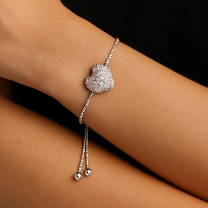 A silver adjustable bracelet with a textured heart pendant, featuring a thin chain and two small metallic beads as part of the closure.