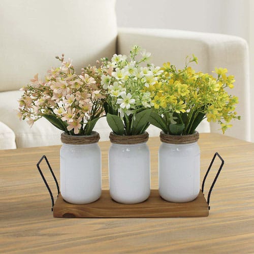 Three white jars with rope accents hold artificial flowers in pink, white, and yellow, placed on a wooden tray with metal handles, on a wooden table.