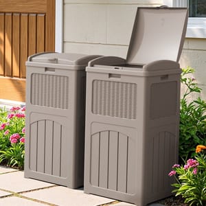 Two beige outdoor storage bins with a wicker texture and hinged lids are placed on a patio next to a flower bed.