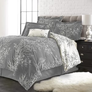 A gray and white bedding set with a nature-inspired branch pattern, featuring a comforter, pillow shams, decorative pillow, and a white throw. A black nightstand with a white lamp and a white fur rug are also visible.