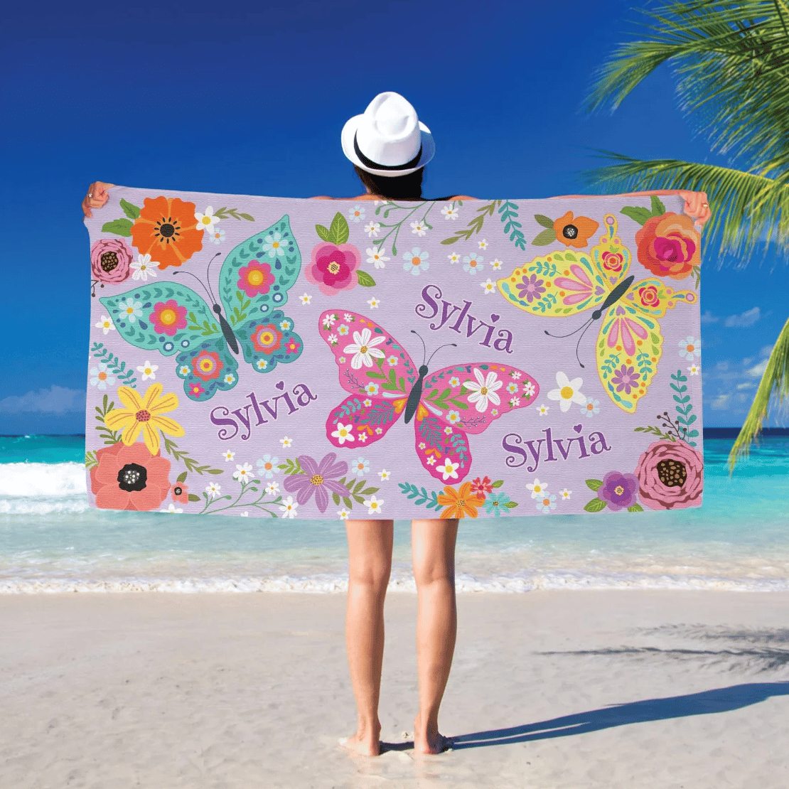 A person at the beach holds a towel decorated with colorful butterflies and flowers, featuring the name \“Sylvia\“ on it.